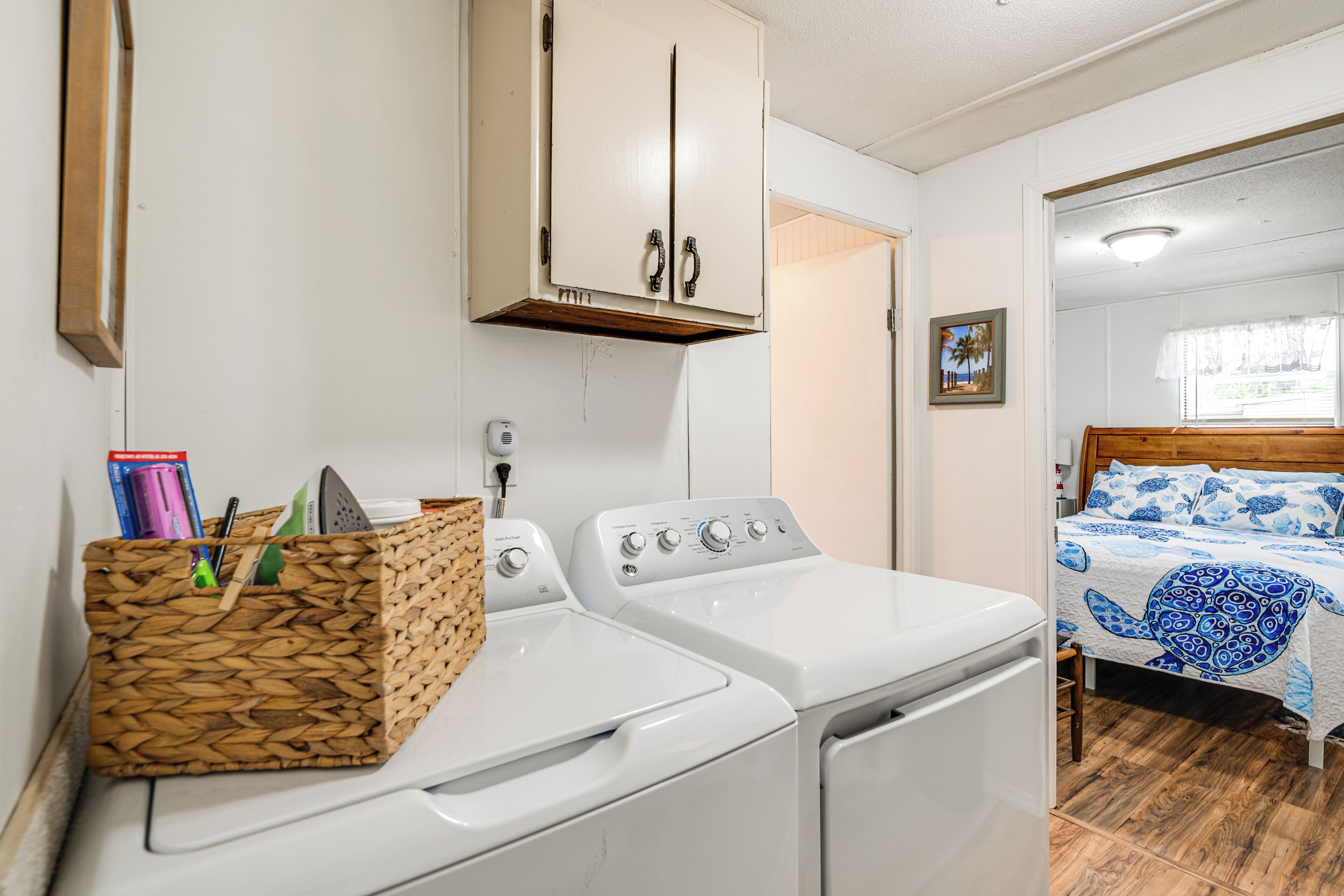 A laundry area with two washing machines, a wooden basket with cleaning supplies, and a bed with a blue and white patterned bedspread.