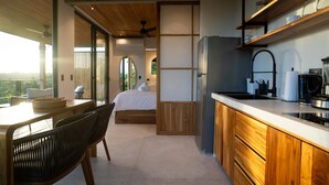 A modern kitchen with wooden cabinets and a built-in refrigerator. There is a dining area with a table and chairs, and a bedroom with a large window and a bed. - w Stylish 1BR w Stunning Balcony Views (Cóbano)
