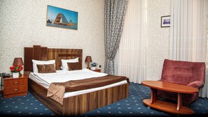 A hotel room with a wooden bed, a red armchair, a small wooden table, a nightstand with a lamp, and a painting on the wall.
