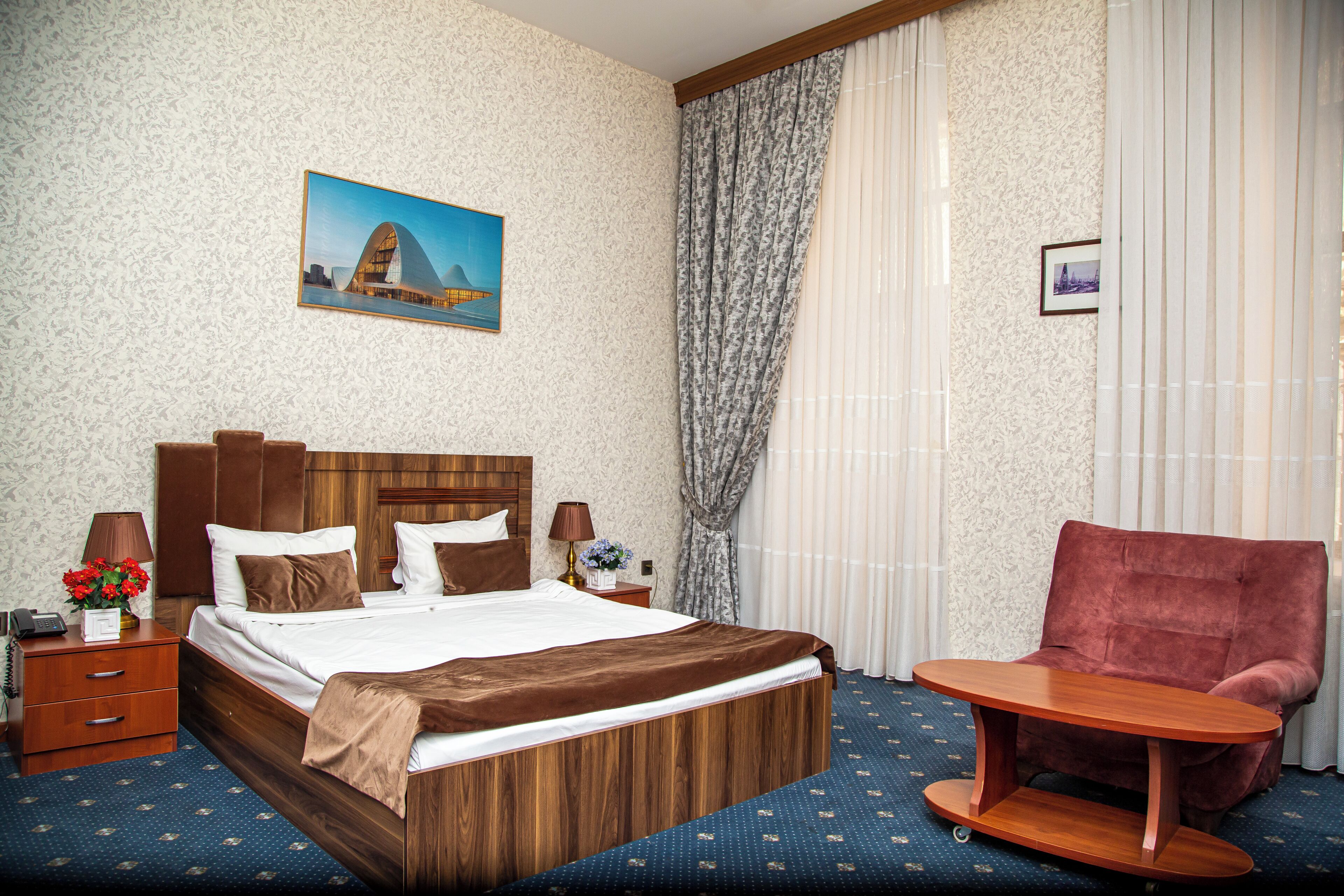 A hotel room with a wooden bed, a small round table, a red armchair, and a painting of a building on the wall.