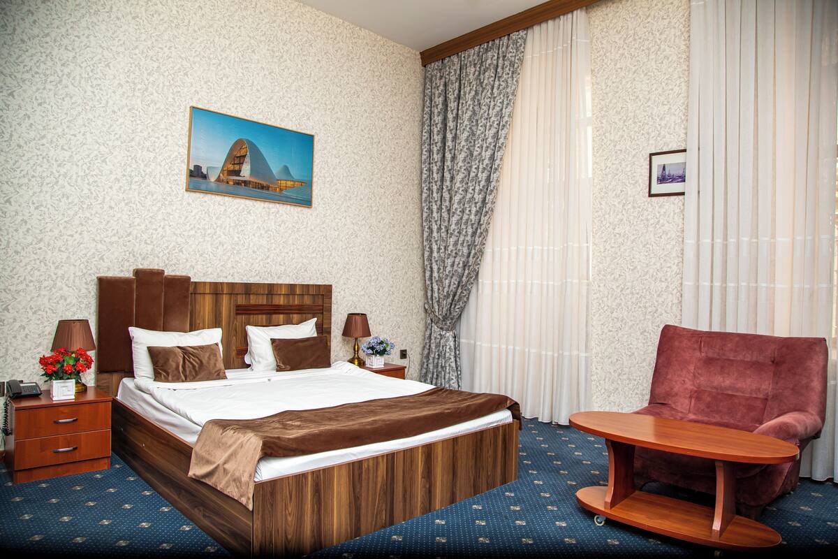 Una camera d'albergo con un letto in legno, una poltrona rossa, un tavolino in legno, un comodino con una lampada e un quadro appeso al muro.