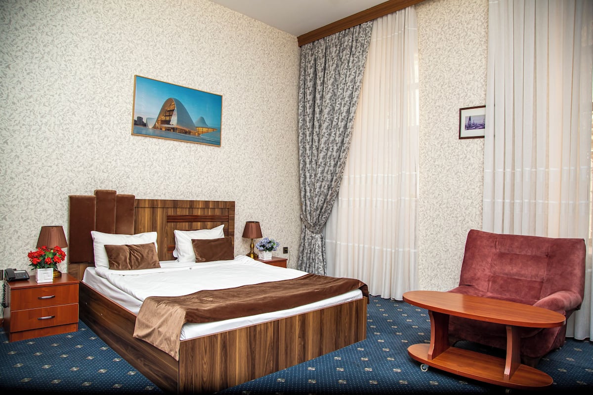 Hotellihuoneessa on puinen sänky, punainen nojatuoli, pieni puinen pöytä, yöpöytä lampun kanssa ja taulu seinällä.