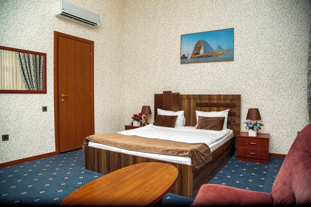 Una camera d'hotel con un letto, una testiera in legno, un comodino con una lampada, una sedia e un condizionatore d'aria a parete.