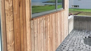 A wooden exterior wall with a large window overlooking a body of water and distant mountains.