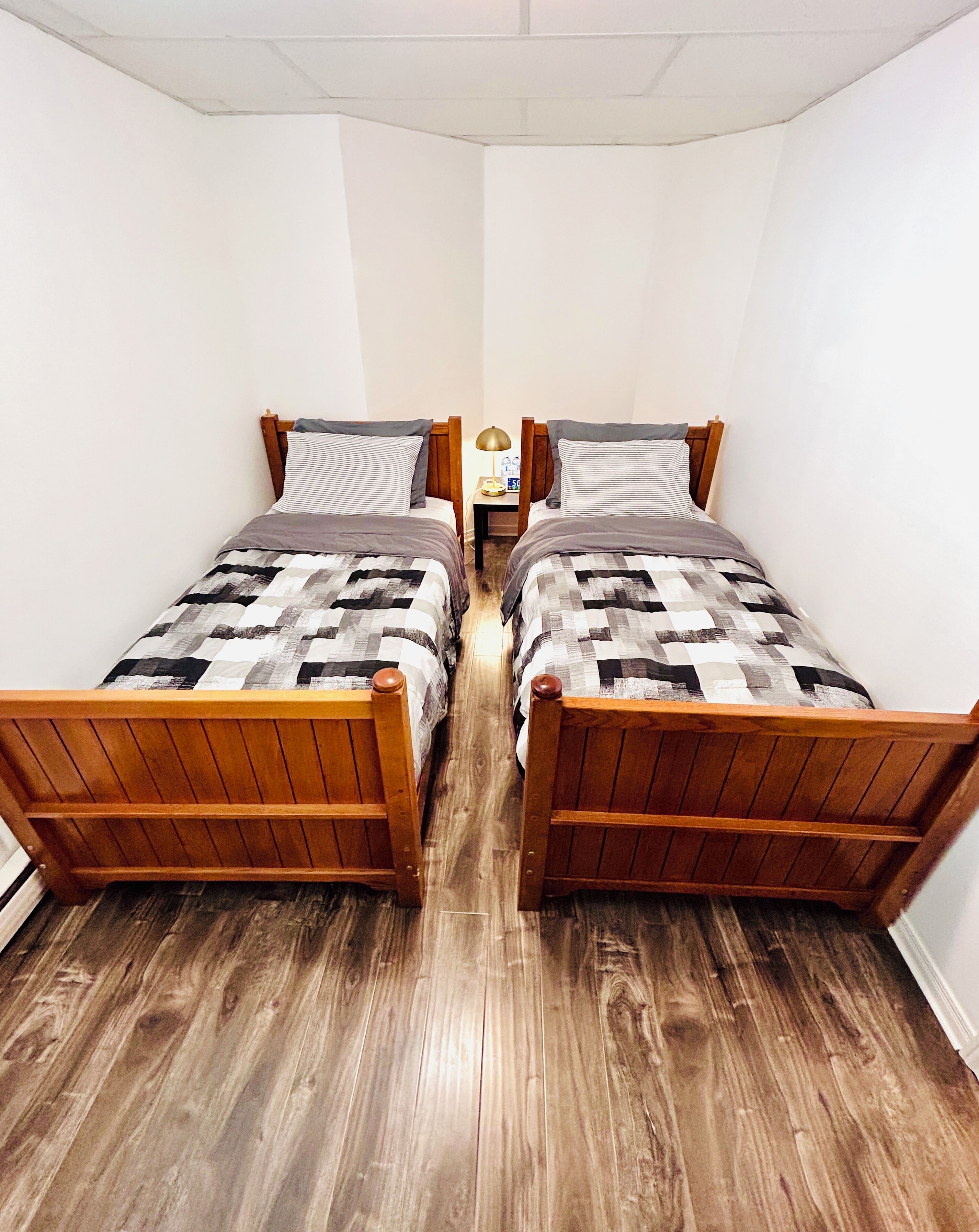 A room with two wooden beds, each with a patterned bedspread and pillows, a bedside table with a lamp, and a wooden floor.