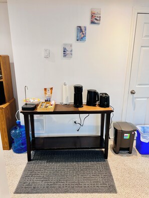 Una habitación con una mesa equipada con cafetera, tostadora y dispensador de agua. Hay un cubo de basura y una jarra de agua azul en el suelo.