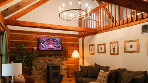 A cozy log cabin interior with a stone fireplace, wooden beams, a television, and a comfortable seating area.