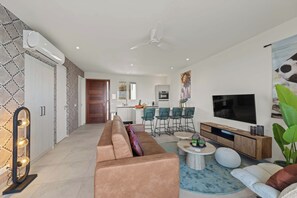 A modern living room with a sofa, coffee table, and TV. There is a kitchen in the background with a dining area. - Caribbean Lounge — Elegant Island Apartment (Blue Bay Golf & Beach Resort)