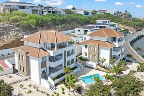 A modern residential complex with multiple two-story buildings, a swimming pool, and a hilly backdrop. - Caribbean Lounge — Elegant Island Apartment (Blue Bay Golf & Beach Resort)