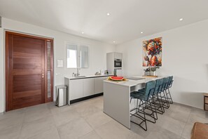 A modern kitchen with a central island, bar stools, a stainless steel trash bin, and a large window. - Caribbean Lounge — Elegant Island Apartment (Blue Bay Golf & Beach Resort)