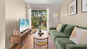Ein Wohnzimmer mit einem grünen Sofa, einem Holztisch, einem Fernseher auf einem Holzgestell und Blick auf einen Garten durch eine Glastür.