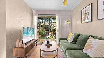 A living room with a green sofa, a wooden coffee table, a TV on a wooden stand, and a view of a garden through a glass door.