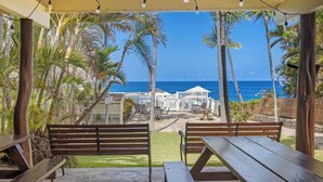 Ein Sitzbereich im Freien mit Holzbänken und -tischen mit Blick auf einen Strand, Palmen und einen klaren, blauen Himmel.