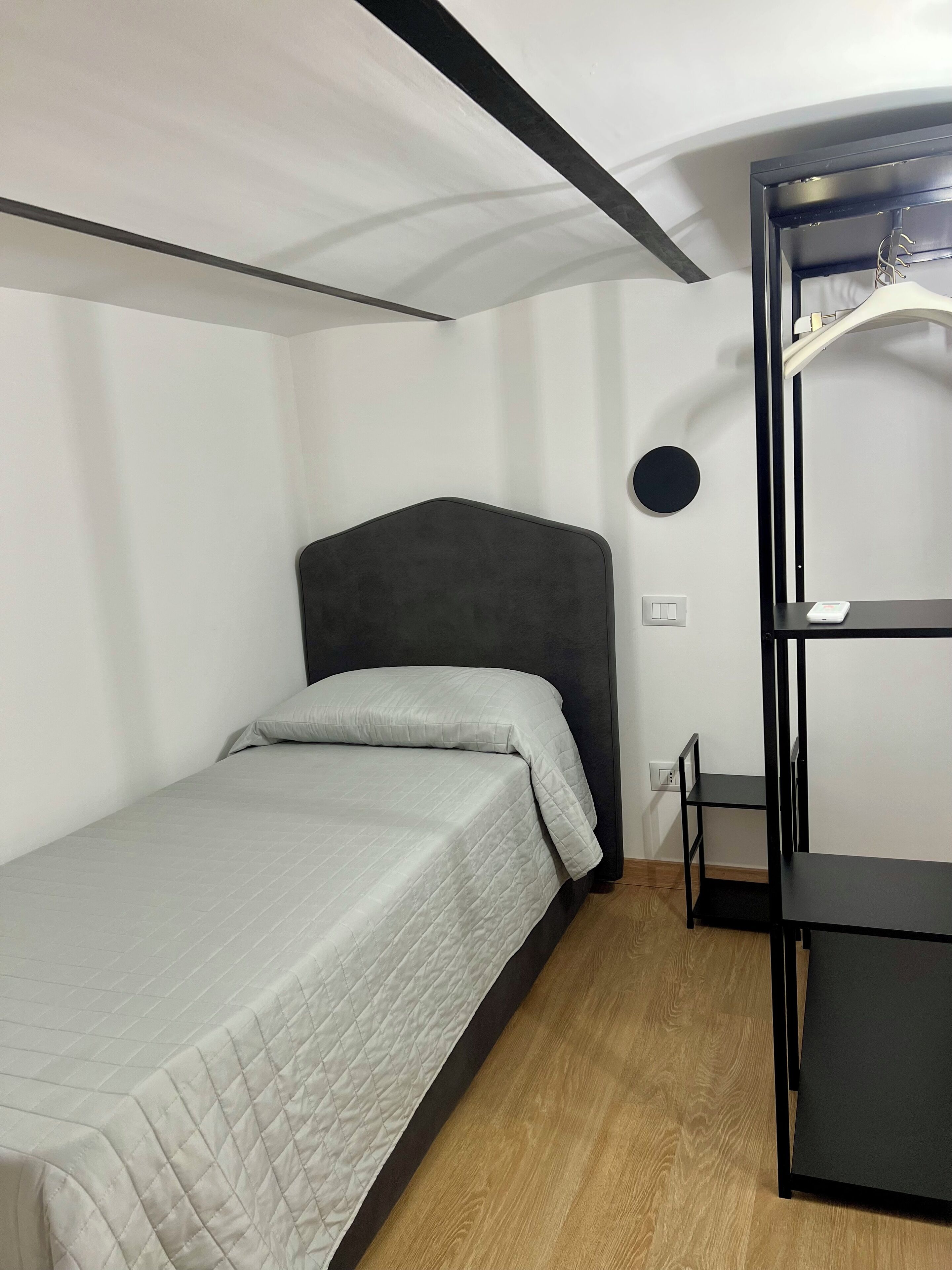 A small, modern room with a single bed, a black headboard, a wooden floor, and a black shelving unit against the wall.