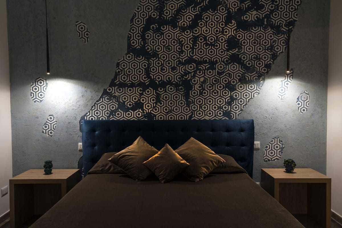 Une chambre à coucher avec une tête de lit capitonnée, un lit avec une literie marron, deux tables de chevet et un mur orné d’un motif géométrique.
