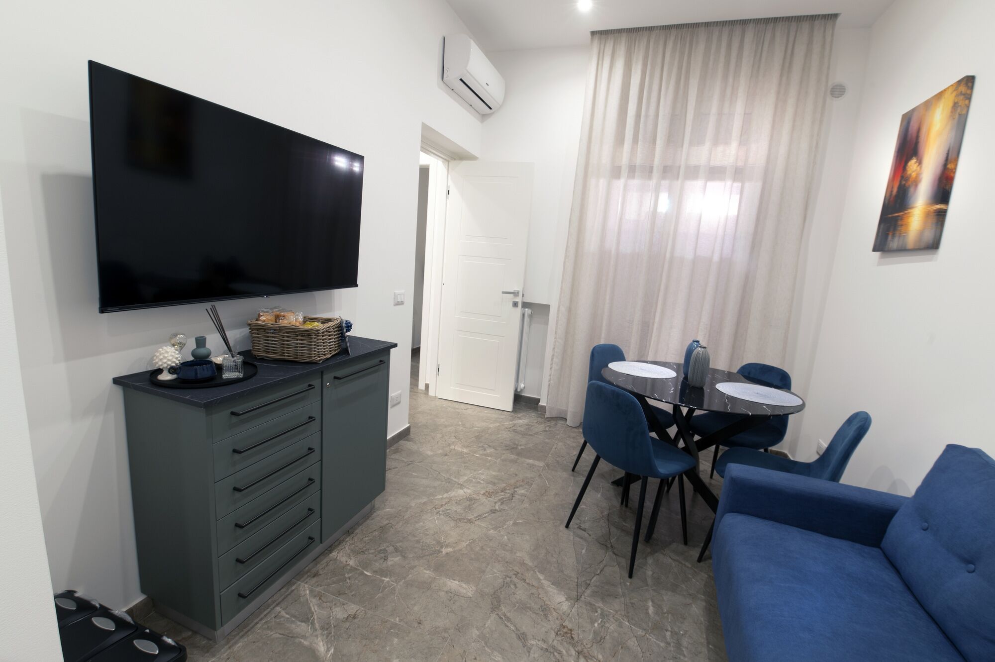 A compact living space with a flat-screen TV mounted on the wall, a dining table with chairs, a sofa, and a small kitchenette area with a cabinet and drawers.