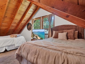 A bedroom with a sloped ceiling, a bed with a beige blanket, a bedside table with a lamp, and a window with a view of a beach.