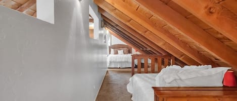 Una habitación con techo inclinado, vigas de madera, una cama con ropa blanca y un objeto rojo sobre la cama.