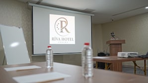 Un salón de conferencias con una pantalla de proyector mostrando el logo del Hotel Riva, un atril, una computadora portátil y dos botellas de agua sobre la mesa.