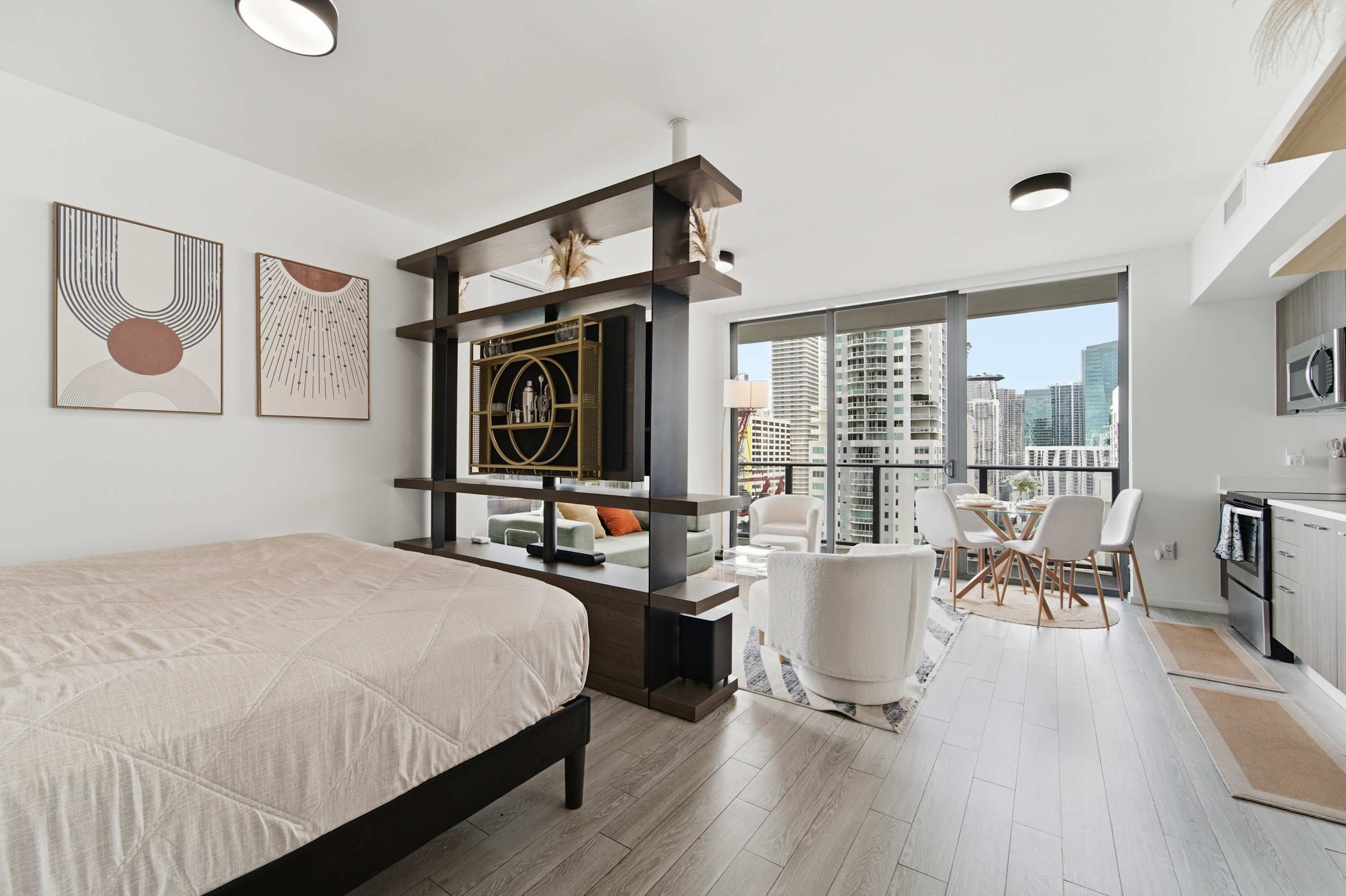 Une chambre d’hôtel moderne comprenant un lit, un coin salon avec une table et des chaises, un coin cuisine et de grandes fenêtres donnant sur la ville.