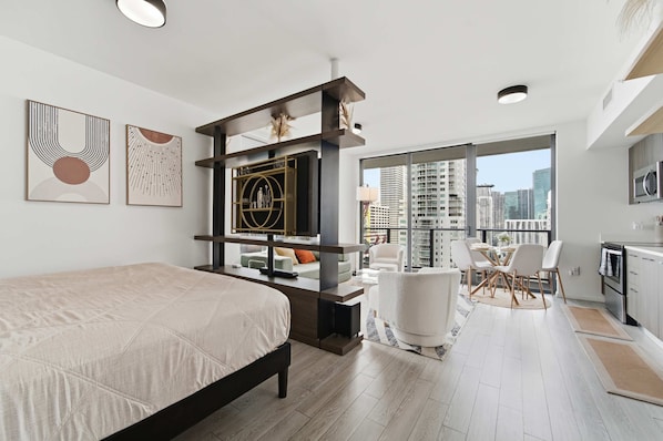 A modern hotel room with a bed, a seating area with a table and chairs, a kitchen area, and large windows offering a city view.