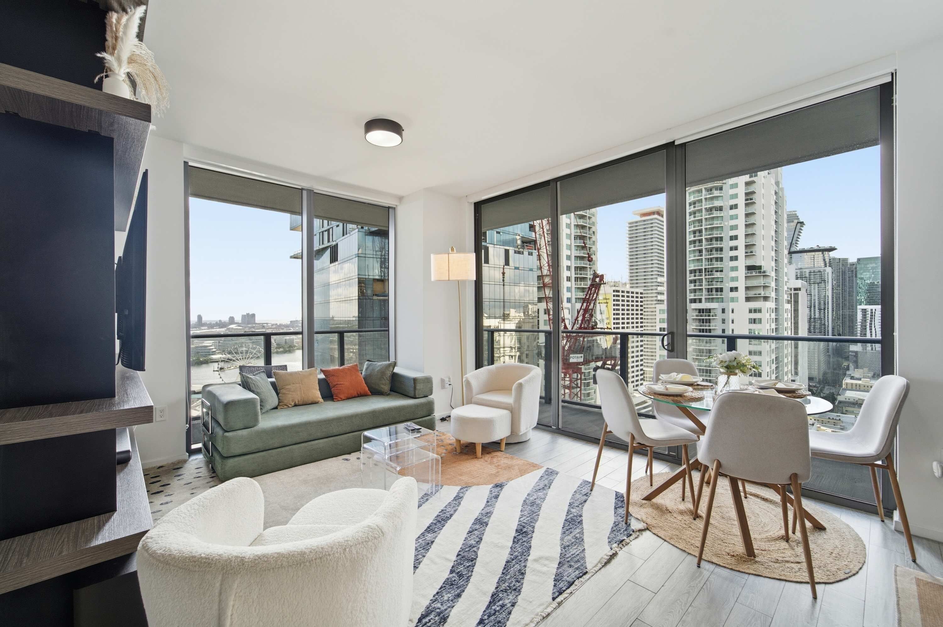 A modern living room with a sofa, chairs, a coffee table, and a dining table with chairs. Large windows offer a view of the cityscape.