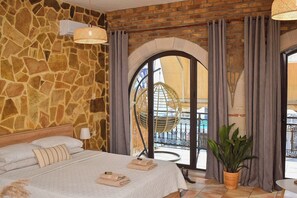 A bedroom with a stone wall, a bed with beige bedding, a hanging chair, and a view of a balcony. - Traditional Cretan house, in the city center! (Chania)