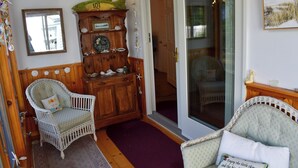 Een gezellige kamer met een rieten stoel, een houten kast met decoratieve objecten, en een glazen deur naar een andere kamer.