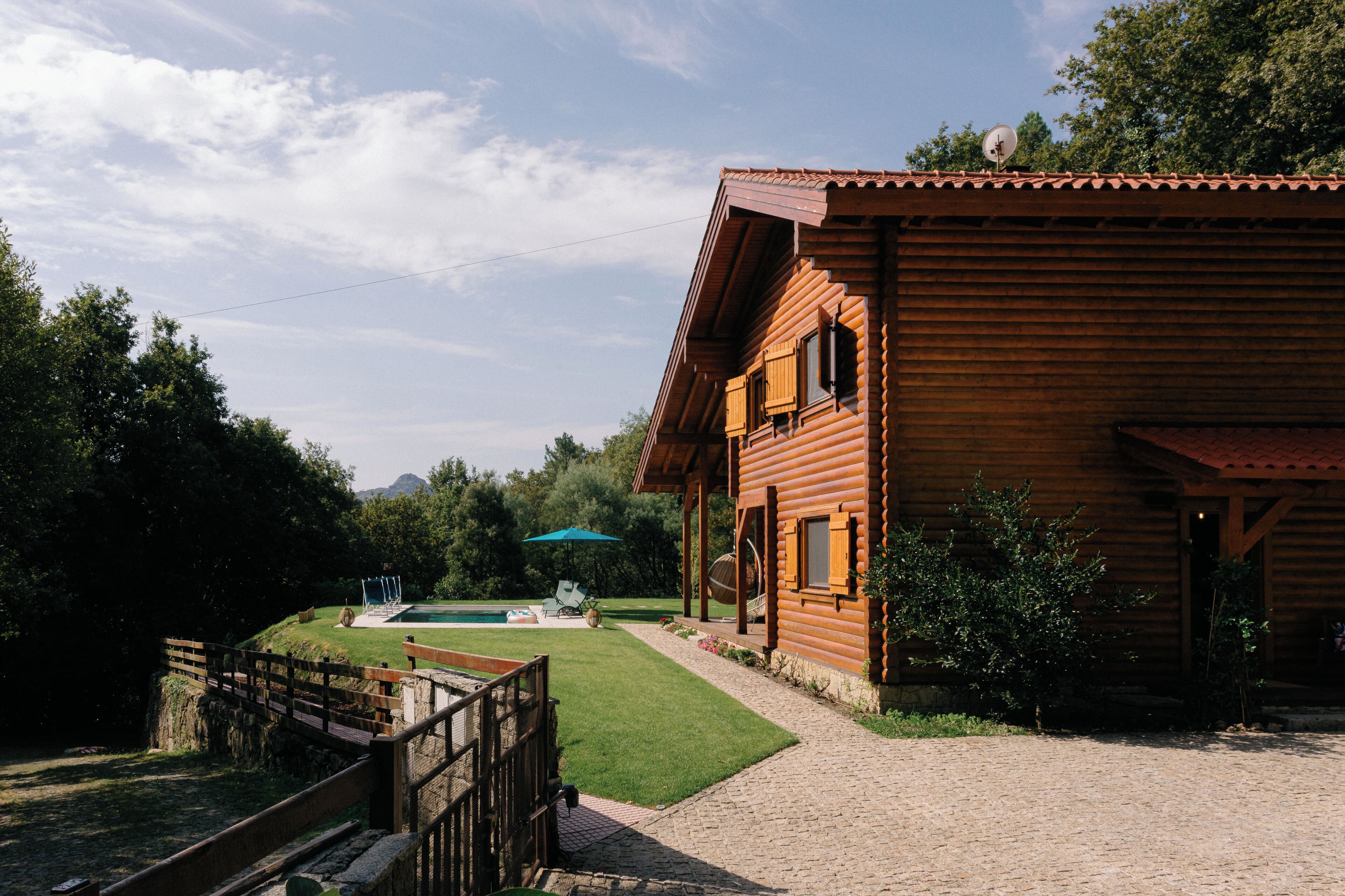 A wooden cabin with a paved driveway, a small lawn, and a bridge.