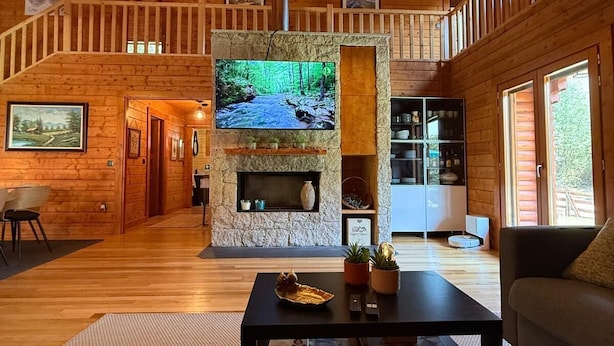 A spacious living room with a stone fireplace, wooden walls, a large TV, and a staircase leading to an upper level. - Gerês Nature House (Terras de Bouro)