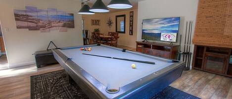 A pool table in a room with a dining area, a TV, and a large window.