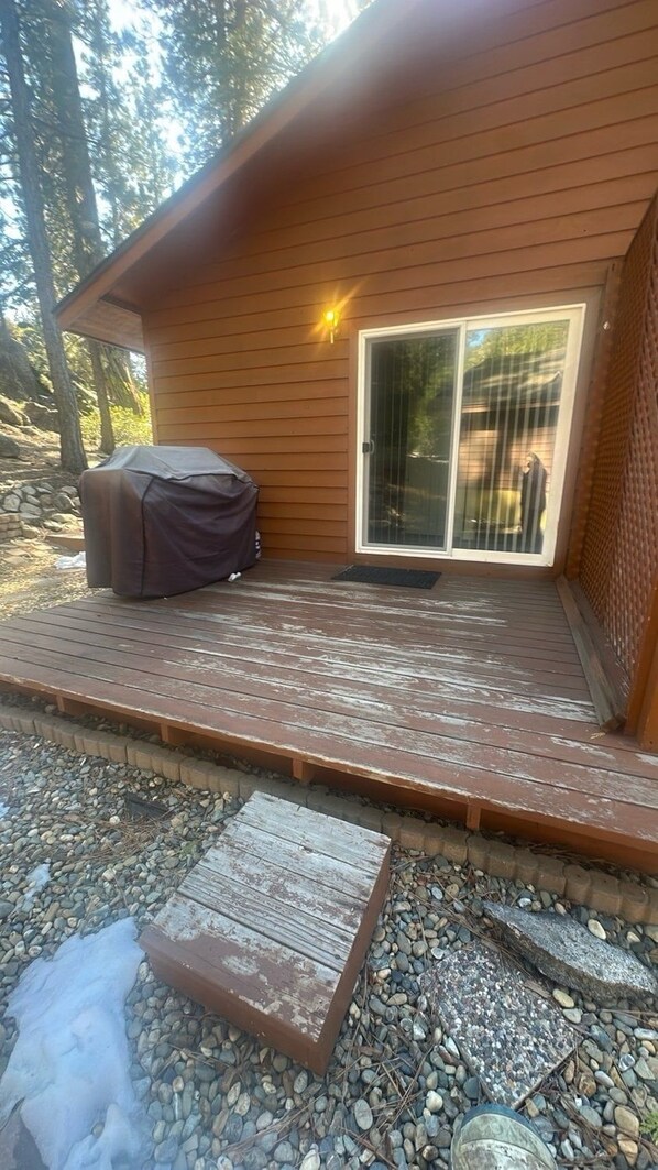 Una terraza de madera con una parrilla cubierta, un ventanal corredero y un sendero de piedra que lleva a la entrada.
