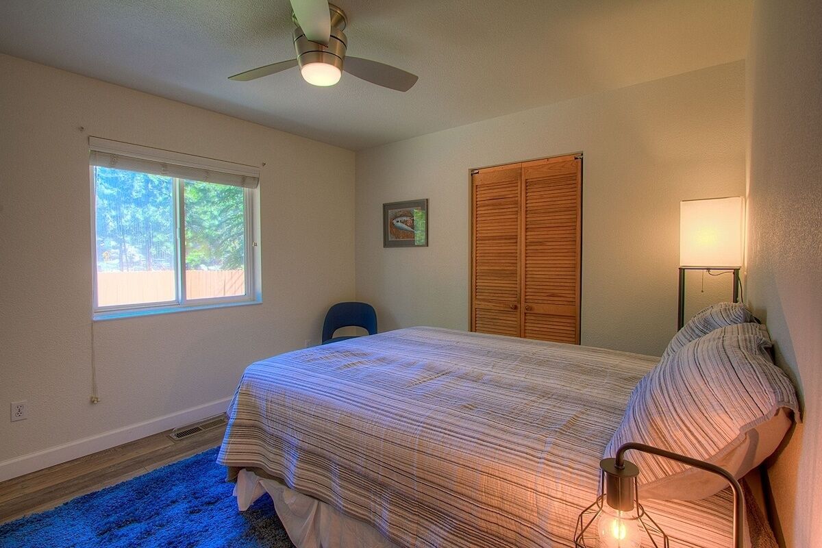 A bedroom with a bed, a ceiling fan, a window, a door, and a nightstand with a lamp.