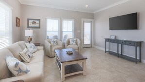 A living room with a sofa, armchairs, a coffee table, and a TV wall-mounted above a grey console. - Cole's Houses 4159-4 4 Bedroom Home (Gulf Shores)
