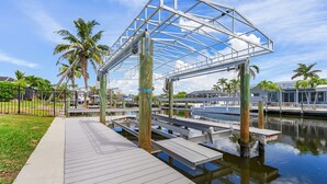 A wooden dock extending into calm water, with a metal structure overhead and palm trees in the background. - 7 Dockside Dream: 3br/3ba Pool Home W/ Water Views! 3 Bedroom Home (Fort Myers Beach)