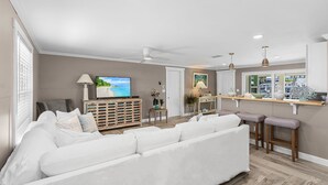 A modern living room with a white sofa, a TV, and a kitchen area with a bar. - 7 Dockside Dream: 3br/3ba Pool Home W/ Water Views! 3 Bedroom Home (Fort Myers Beach)