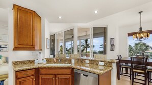 A kitchen with granite countertops, wooden cabinets, a dishwasher, and a dining area with a table and chairs. - Waterside #835: Bayscape Bliss 3br/2ba Condo W/ Pool! 3 Bedroom Condo (Fort Myers Beach)