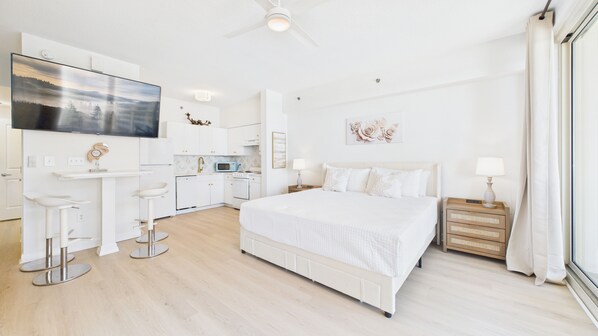 A modern hotel room with a large bed, a TV, a kitchenette, and a bathroom.