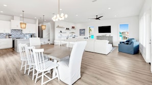 Une cuisine moderne avec des meubles blancs, une table à manger en bois et des chaises blanches. On y trouve un grand îlot central doté d’un évier et d’un four encastré.