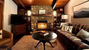 Ein gemütliches Wohnzimmer mit Kamin aus Stein, Holzbalkendecke, Fernseher und einem karierten Sofa.