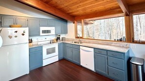 Eine moderne Küche mit Holzbalkendecke, blauen Schränken, einem weißen Kühlschrank und einem großen Fenster mit Blick auf Bäume.