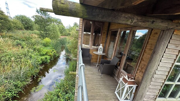A wooden deck with a view of a river, a small table with a lantern, and wicker furniture. - Huge open plan Log Cabin in Weston, Newark hot tub WiFi wood stove pets parking (Weston)