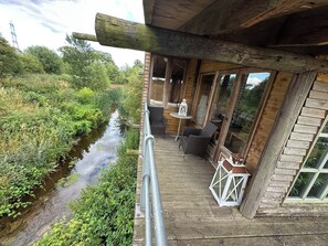 A wooden deck with a view of a river, a small table with a lantern, and wicker furniture. - Riverside wood lodge in Weston, Newark hot tub WiFi wood stove pets parking (Weston)