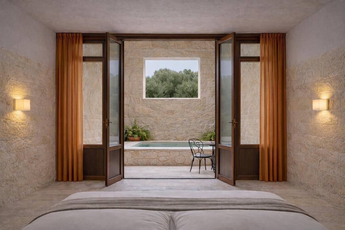 Ein Schlafzimmer mit einem großen Bett, Blick auf eine Steinmauer, einem Fenster und einer kleinen Sitzgelegenheit im Freien mit Tisch und Stühlen.