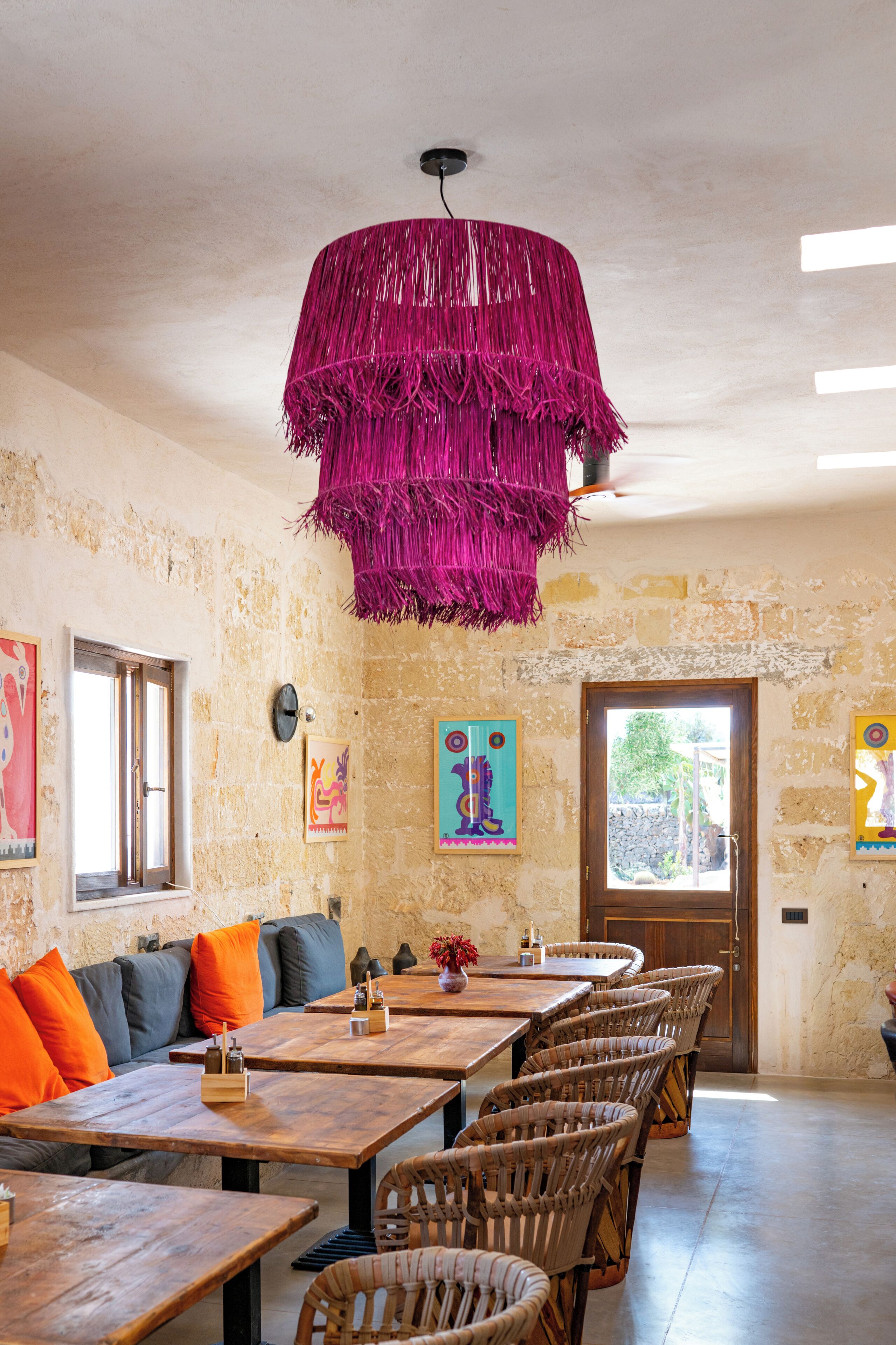 A dining area with wooden tables and wicker chairs, a stone wall, and a vibrant pink hanging light fixture.