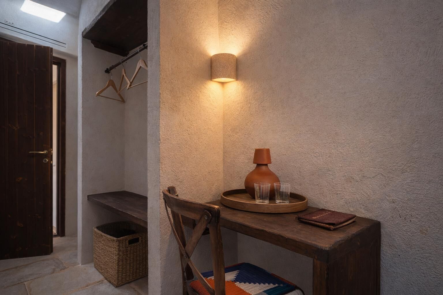 A small, cozy room with a wooden bench, a wall-mounted light, a woven basket, and a rustic vase.