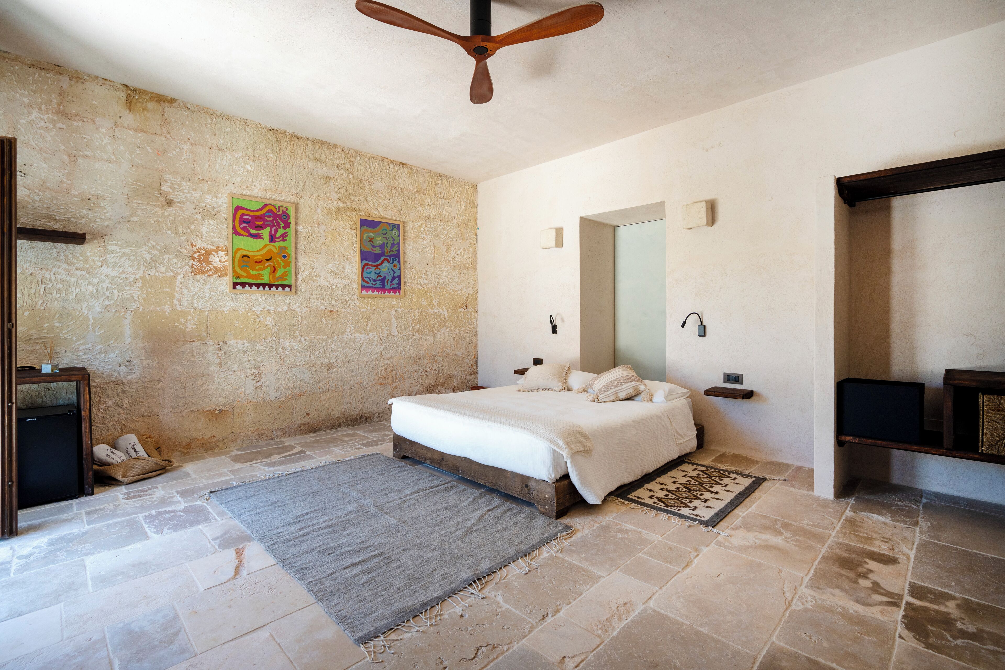 Un dormitorio con cama, ventilador de techo, suelo de piedra y dos cuadros de colores en las paredes.
