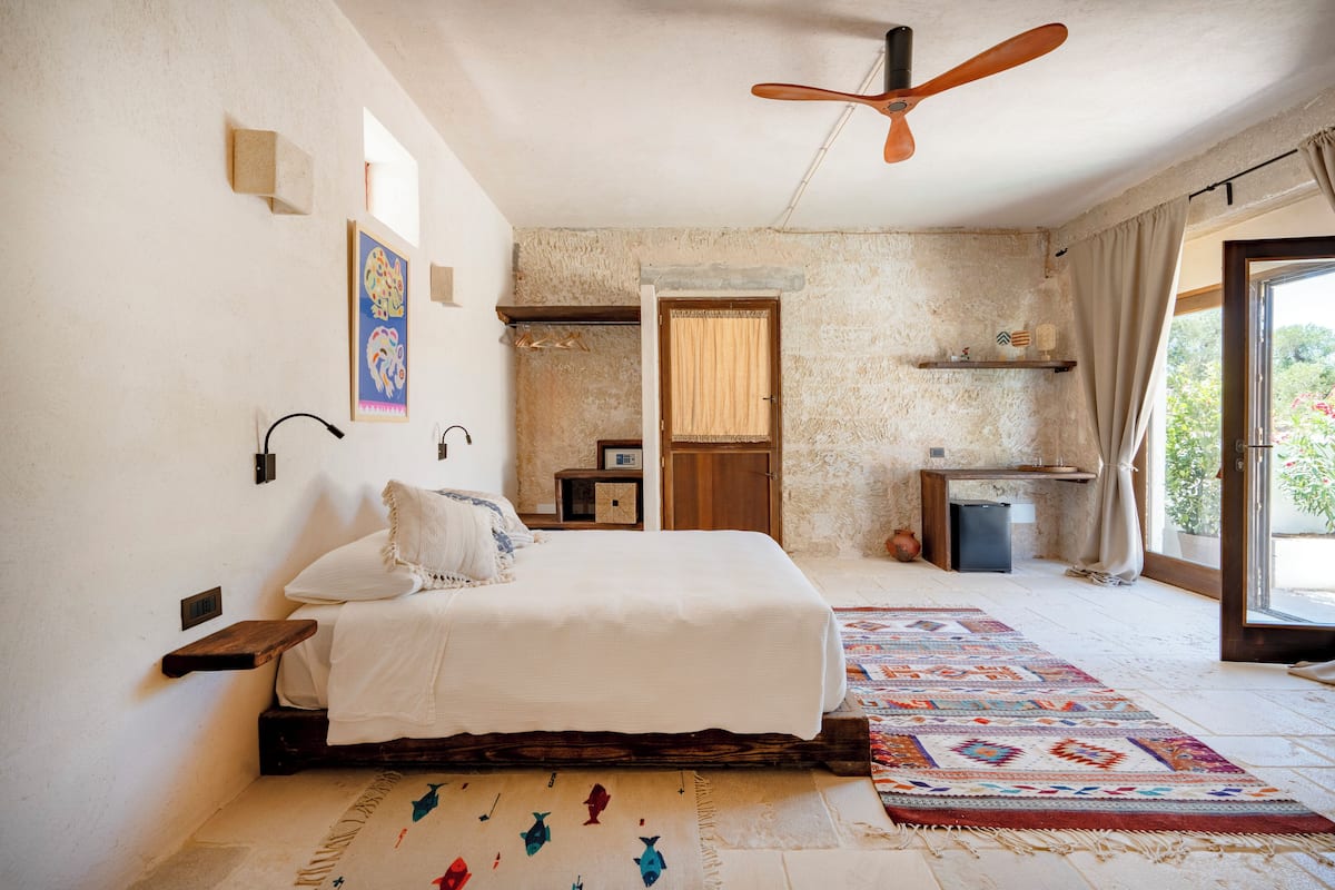 Ein Schlafzimmer mit Bett, Deckenventilator, Teppich, einem kleinen Tisch und einem Fenster mit Vorhängen.