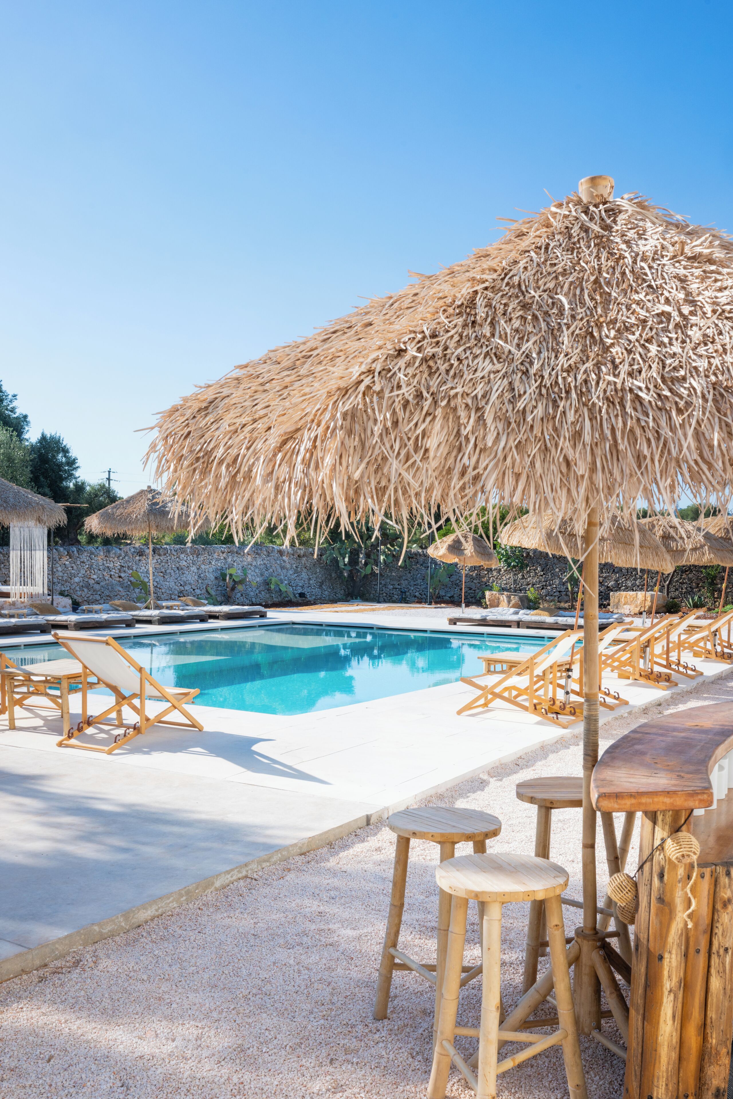 Un espace piscine agrémenté d’un parasol en chaume, de chaises longues en bois et d’un mur de pierre en arrière-plan.