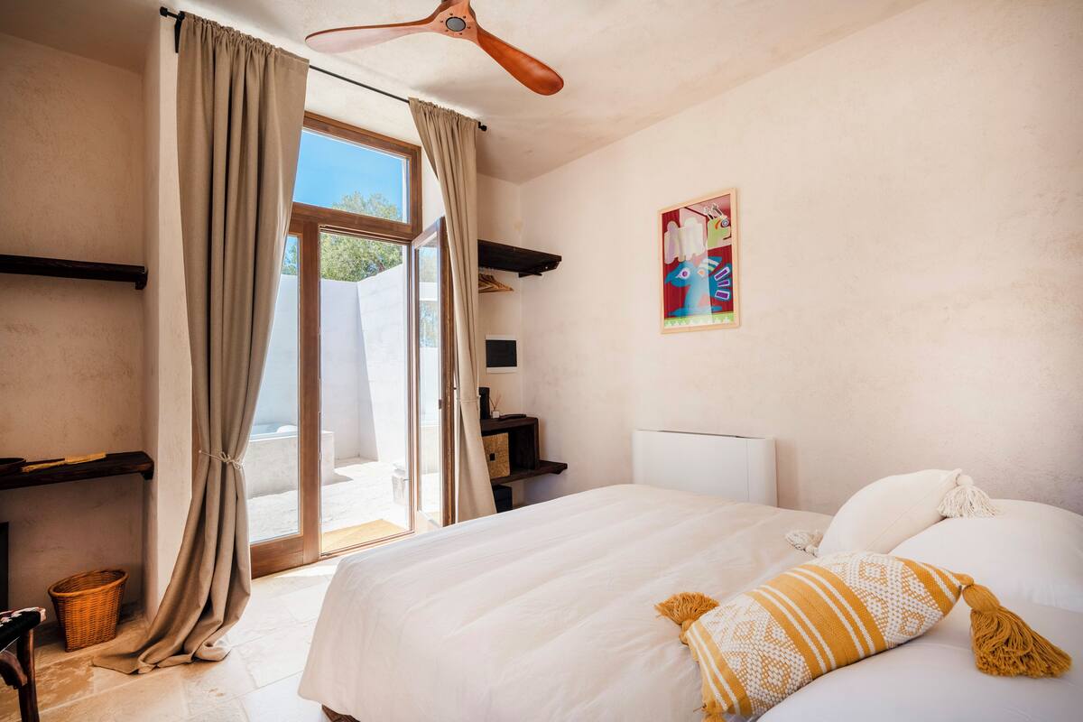 Ein Schlafzimmer mit einem großen Bett, einem Deckenventilator, einem Fenster mit Vorhängen, einem an der Wand montierten Regal und einem farbenfrohen Gemälde.